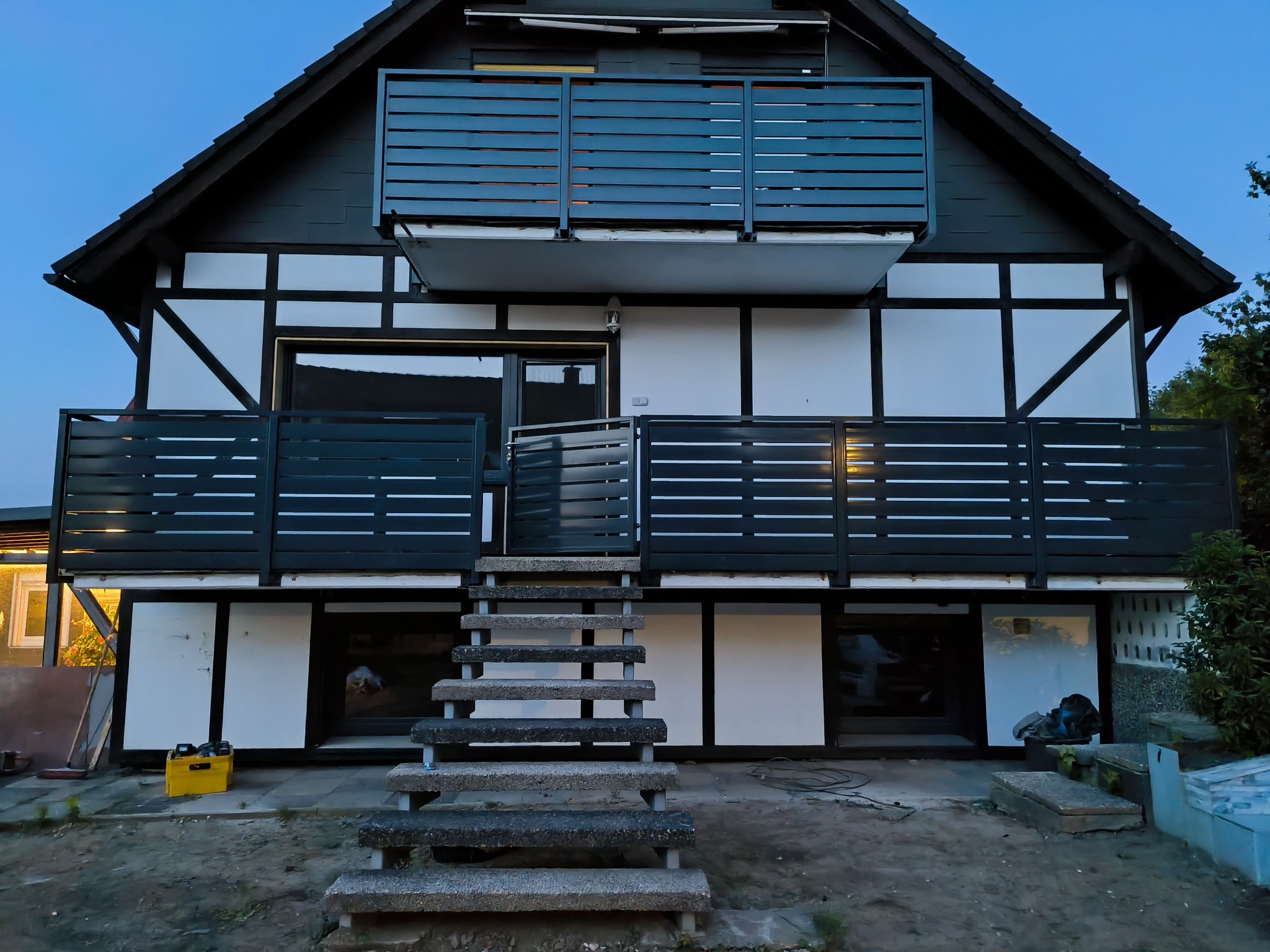 Dom w stylu szachulcowym z nowoczesnymi, metalowymi balustradami balkonowymi w kolorze grafitowym. Widoczne betonowe schody wejściowe i elementy wyposażenia.