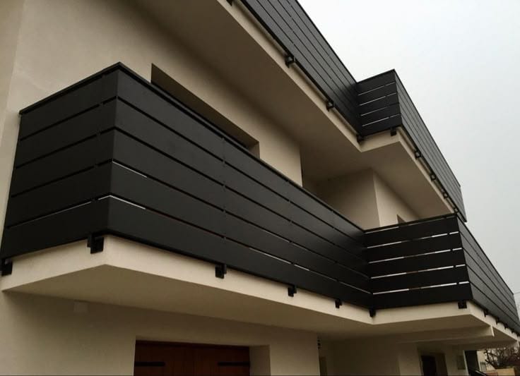 Nowoczesna balustrada balkonowa z poziomych, czarnych paneli, zamontowana na jasnym budynku. Minimalistyczny design, widok z dołu, pochmurne niebo w tle.