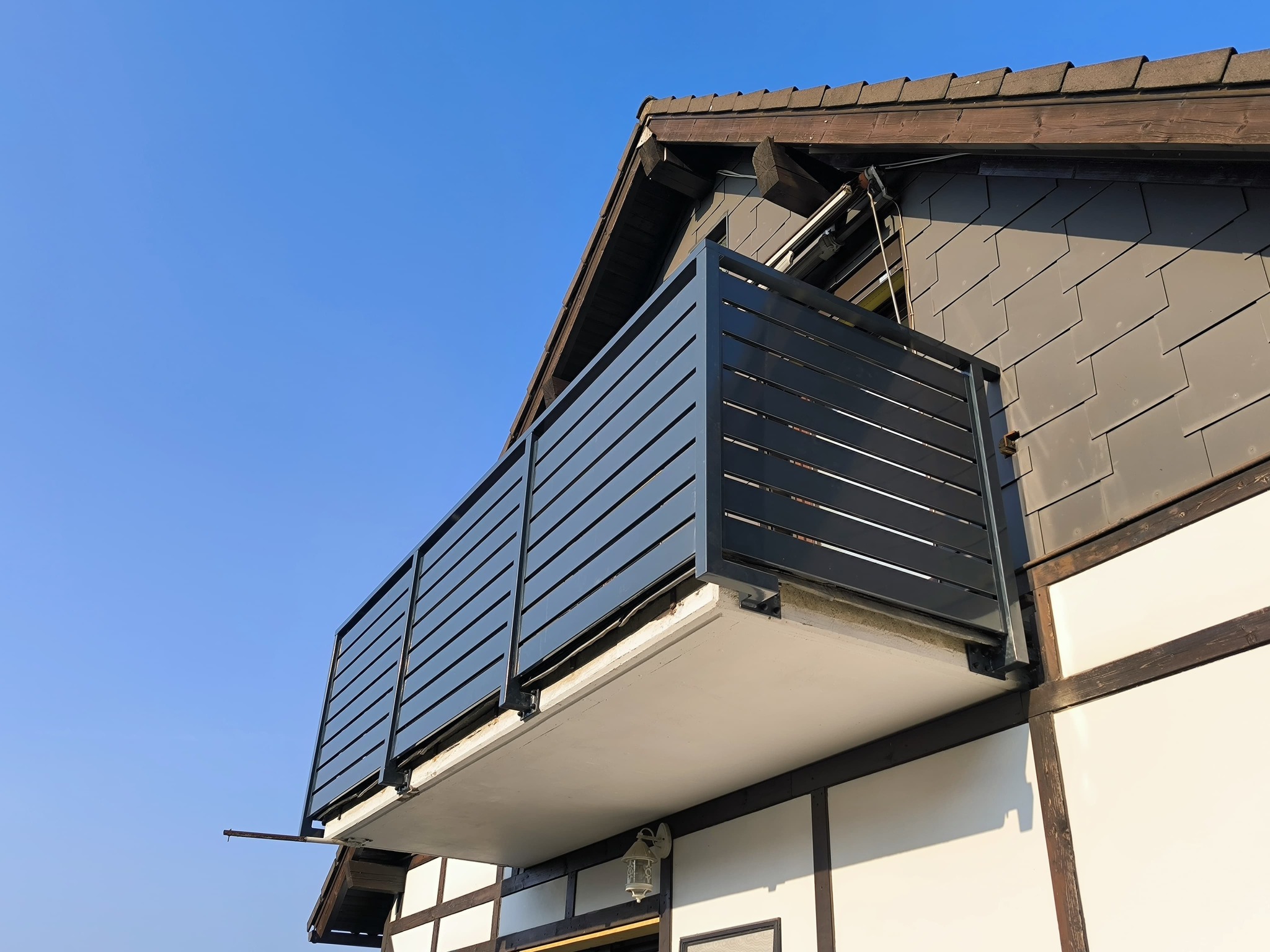 Nowoczesna, pozioma balustrada balkonowa w kolorze antracytowym, zamontowana na balkonie budynku z elementami muru pruskiego, widziana z dołu na tle błękitnego nieba.