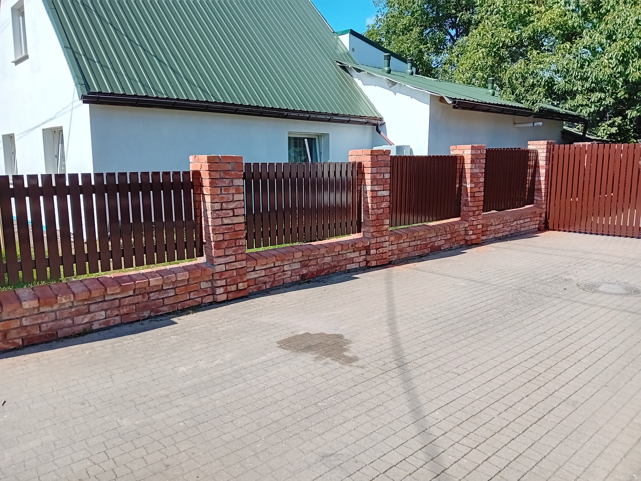 Brązowe, drewniane ogrodzenie z ceglanymi słupkami na tle białego domu z zielonym dachem, widziane z poziomu chodnika z kostki brukowej.