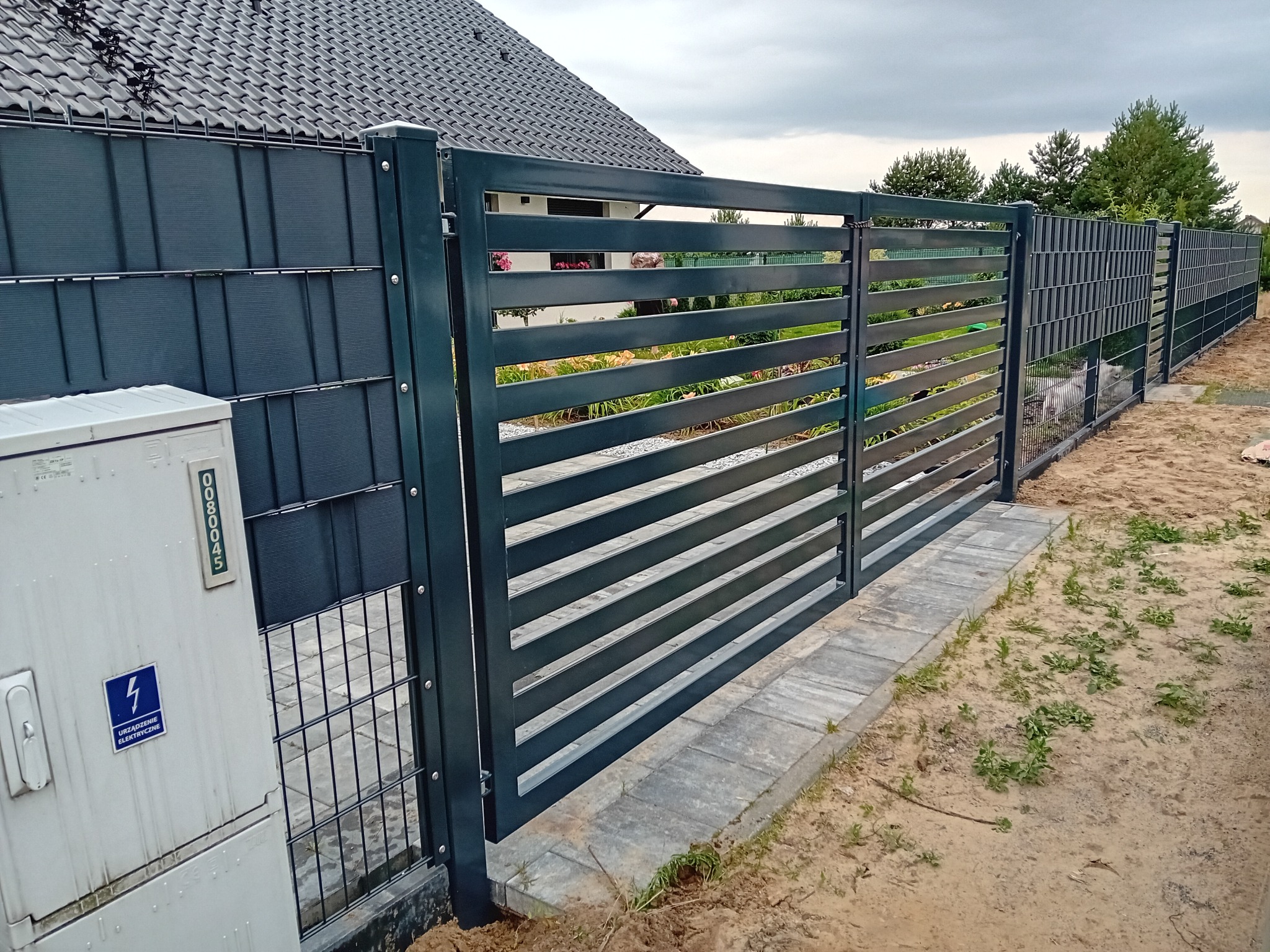 Nowoczesna, pozioma brama metalowa w kolorze grafitowym, z widocznymi elementami ogrodzenia panelowego, na tle domu z szarą dachówką. Widoczna skrzynka elektryczna.