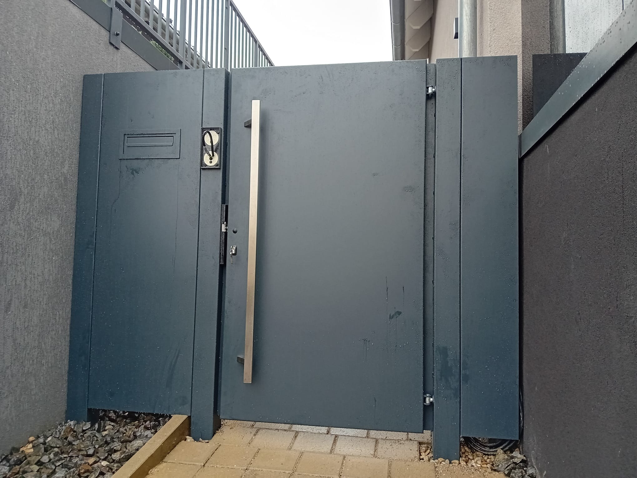 Nowoczesna, grafitowa brama wejściowa z długim, srebrnym uchwytem i skrzynką na listy, zamontowana w Dębnie. Widoczne kamienie ozdobne u podstawy.