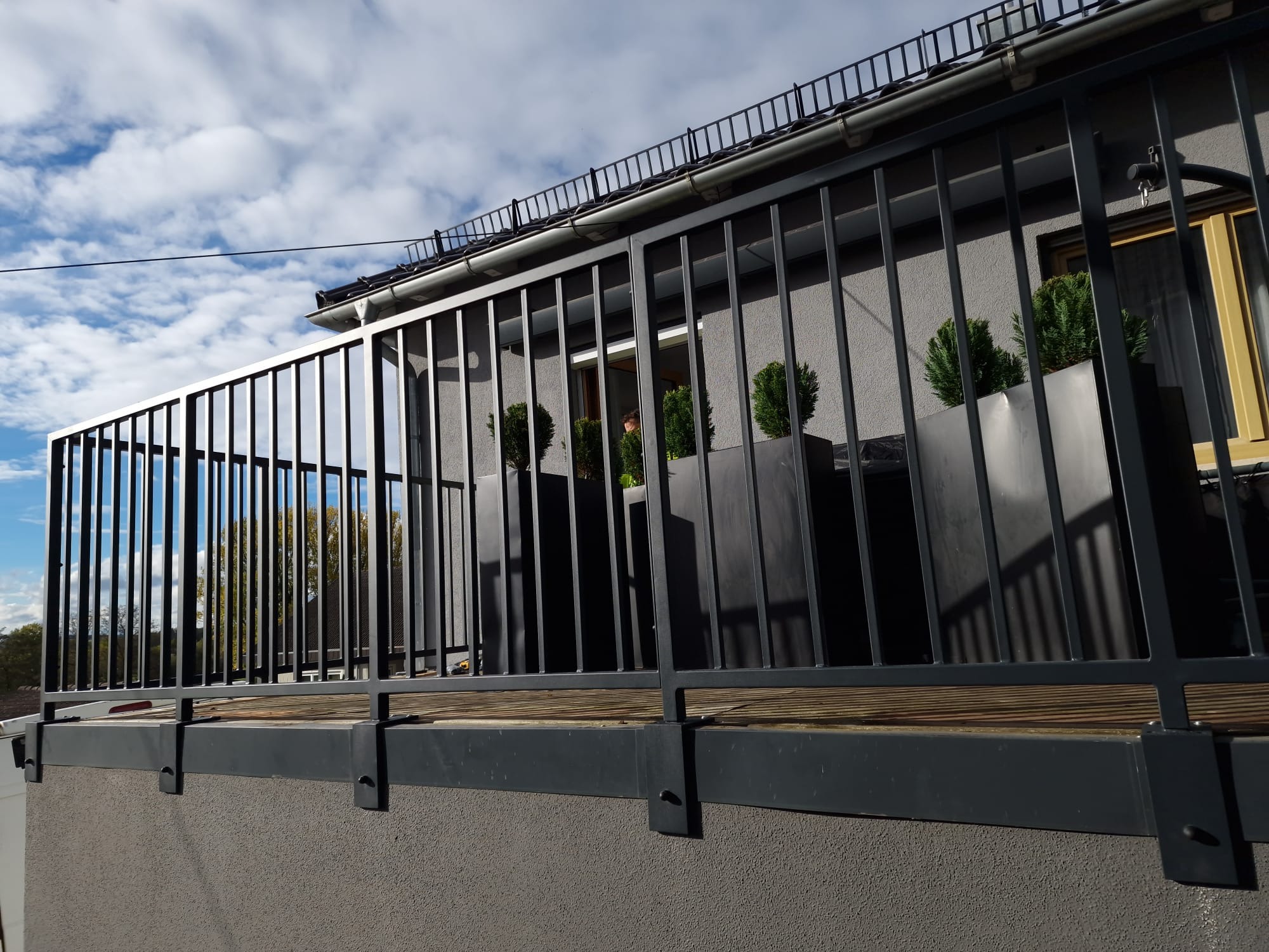 Nowoczesna, grafitowa balustrada balkonowa z pionowymi szczeblami, zamontowana na szarym balkonie z donicami i zielonymi roślinami. Widok z dołu na tle błękitnego nieba.