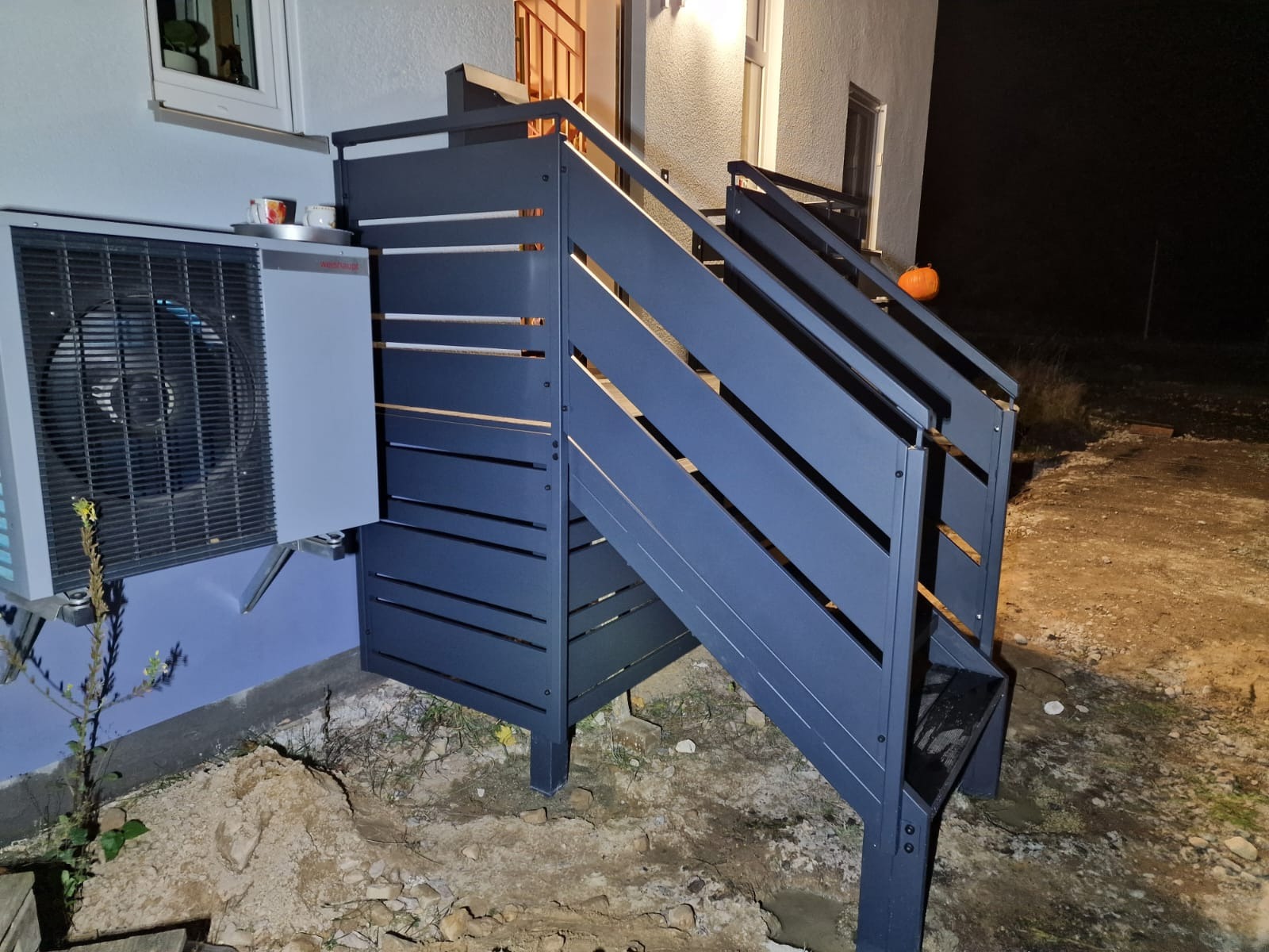 Nowoczesna, grafitowa balustrada schodowa z poziomymi szczeblami, zamontowana przy budynku. Widoczna pompa ciepła i dynia na schodach. Nocne oświetlenie.