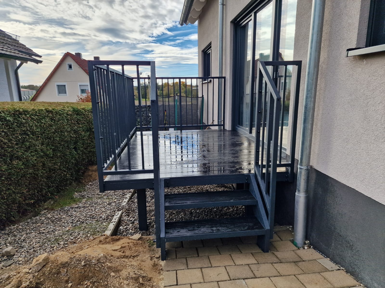 Nowoczesna, metalowa balustrada schodowa w kolorze grafitowym, prowadząca na drewniany taras przy domu. Widoczne odbicia w mokrej nawierzchni tarasu i schodów.