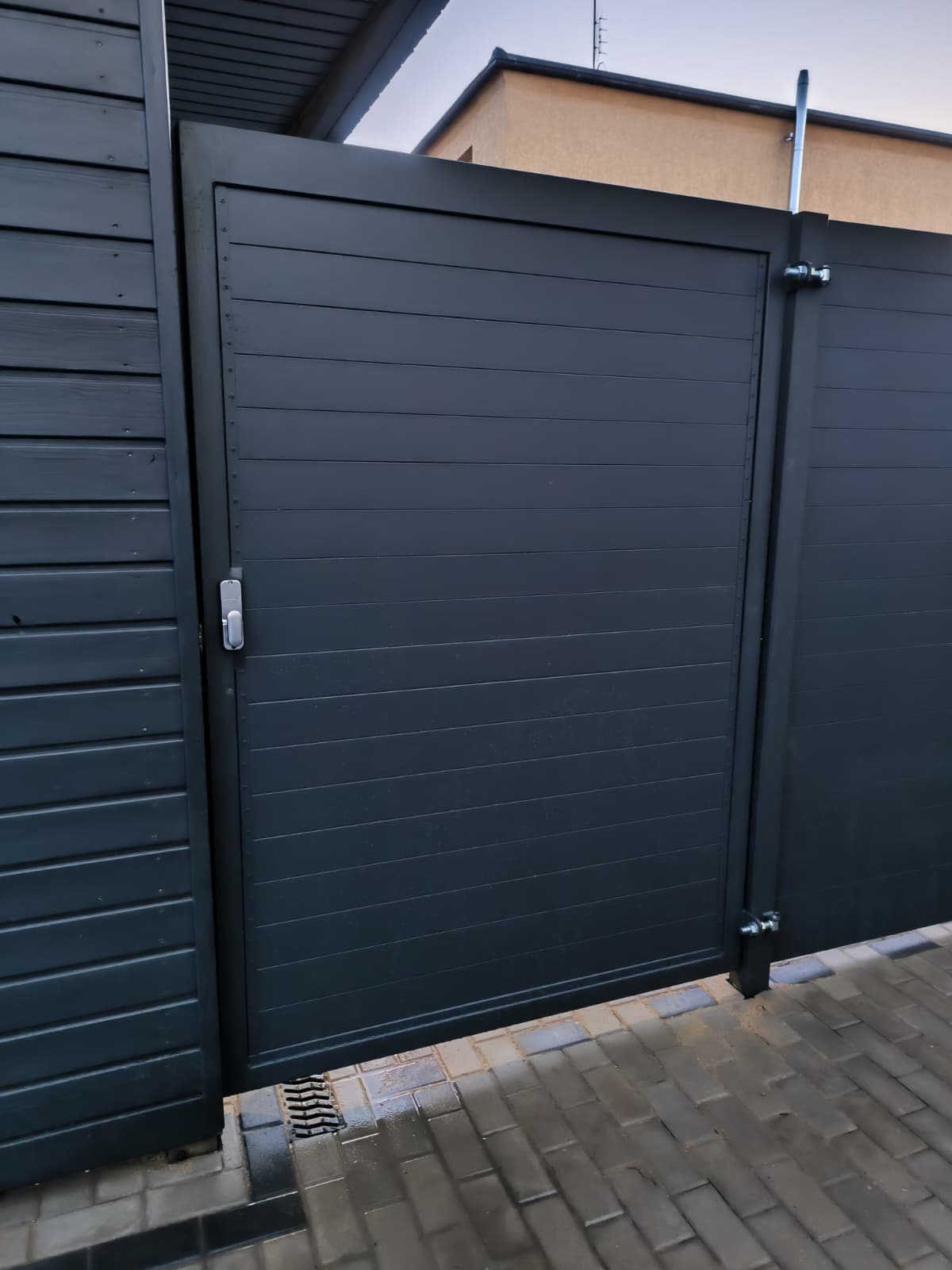 Nowoczesna, grafitowa brama wjazdowa z poziomymi panelami, zamontowana przy drewnianym ogrodzeniu. Widoczny zamek i elementy mocujące. Kostka brukowa na podjeździe.