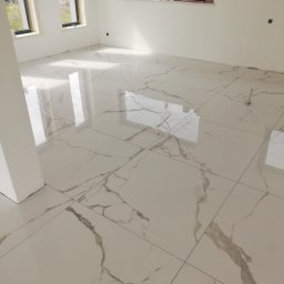 Pro-job - Wyłożona jasnymi, błyszczącymi płytkami podłoga w nowym, pustym pomieszczeniu z trzema oknami i białymi ścianami. Płytki imitują marmur z szarymi żyłami.