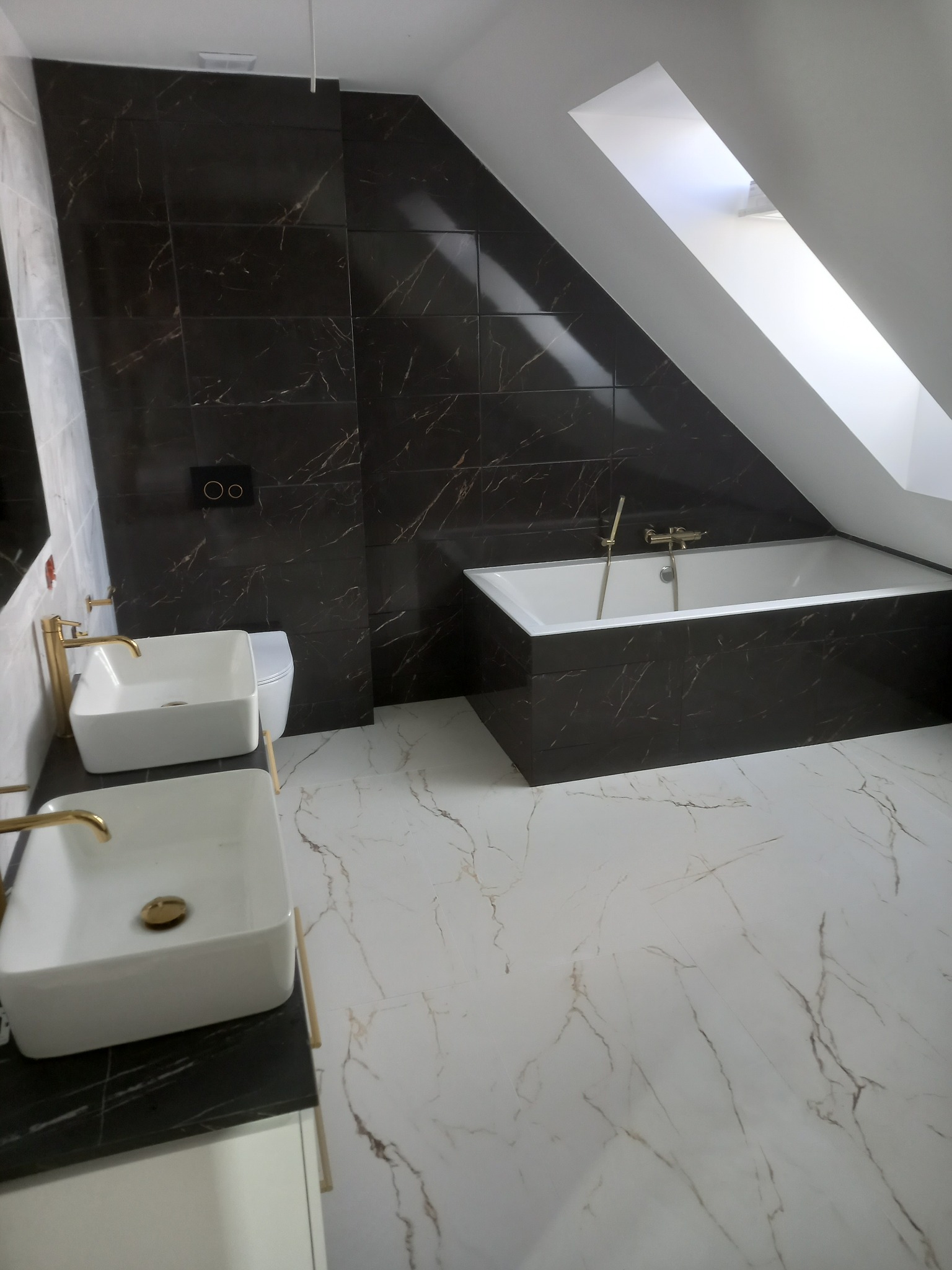 Nowoczesna łazienka na poddaszu z wanną obudowaną płytkami imitującymi marmur, dwiema białymi umywalkami nablatowymi z złotą armaturą i czarną płytką ścienną z marmurowym wzorem.
