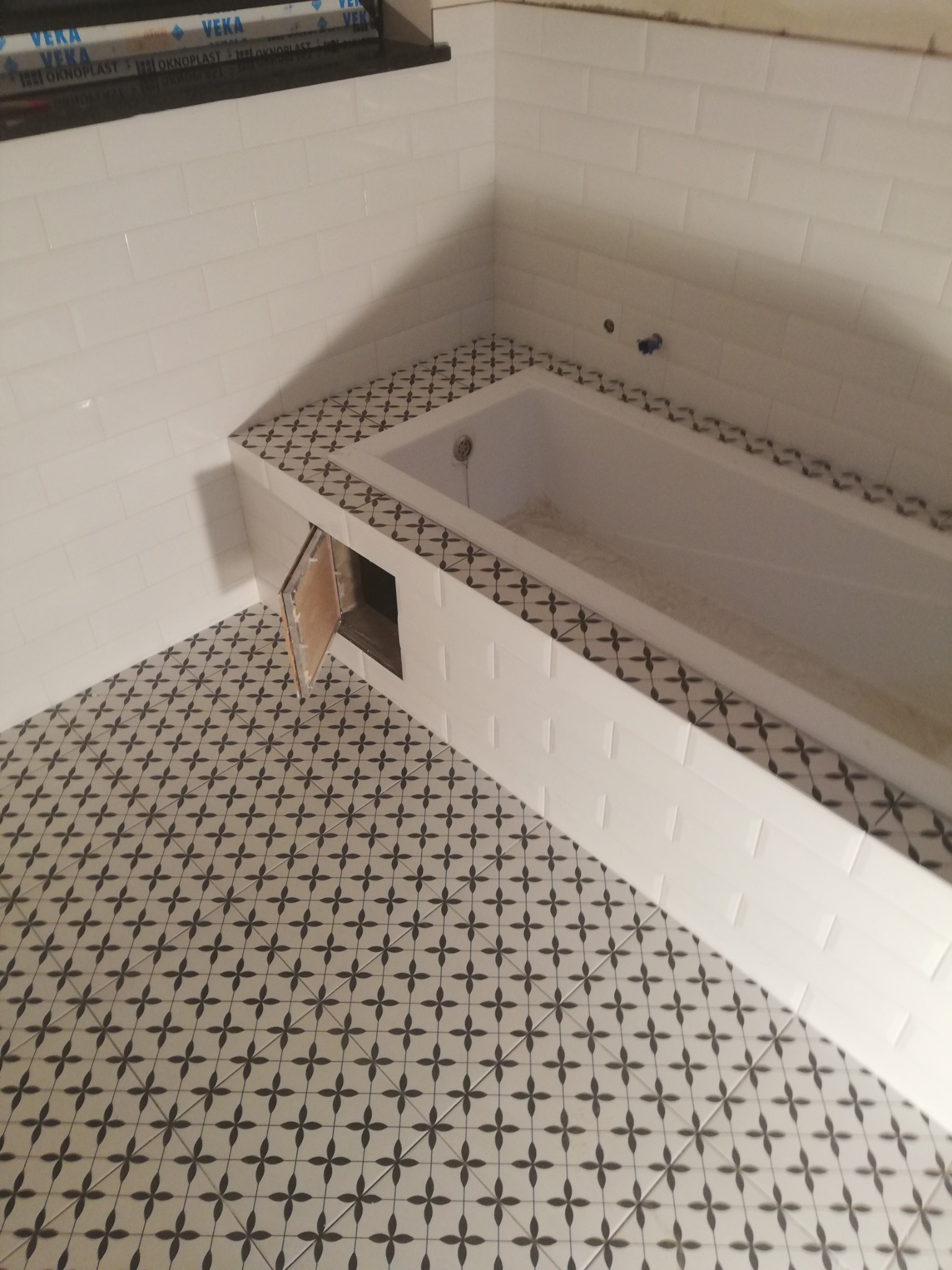 Wykończona łazienka z białą wanną obudowaną białymi płytkami, obrzeżem z płytek dekoracyjnych i podłogą w czarno-białą mozaikę; widoczny otwór rewizyjny.