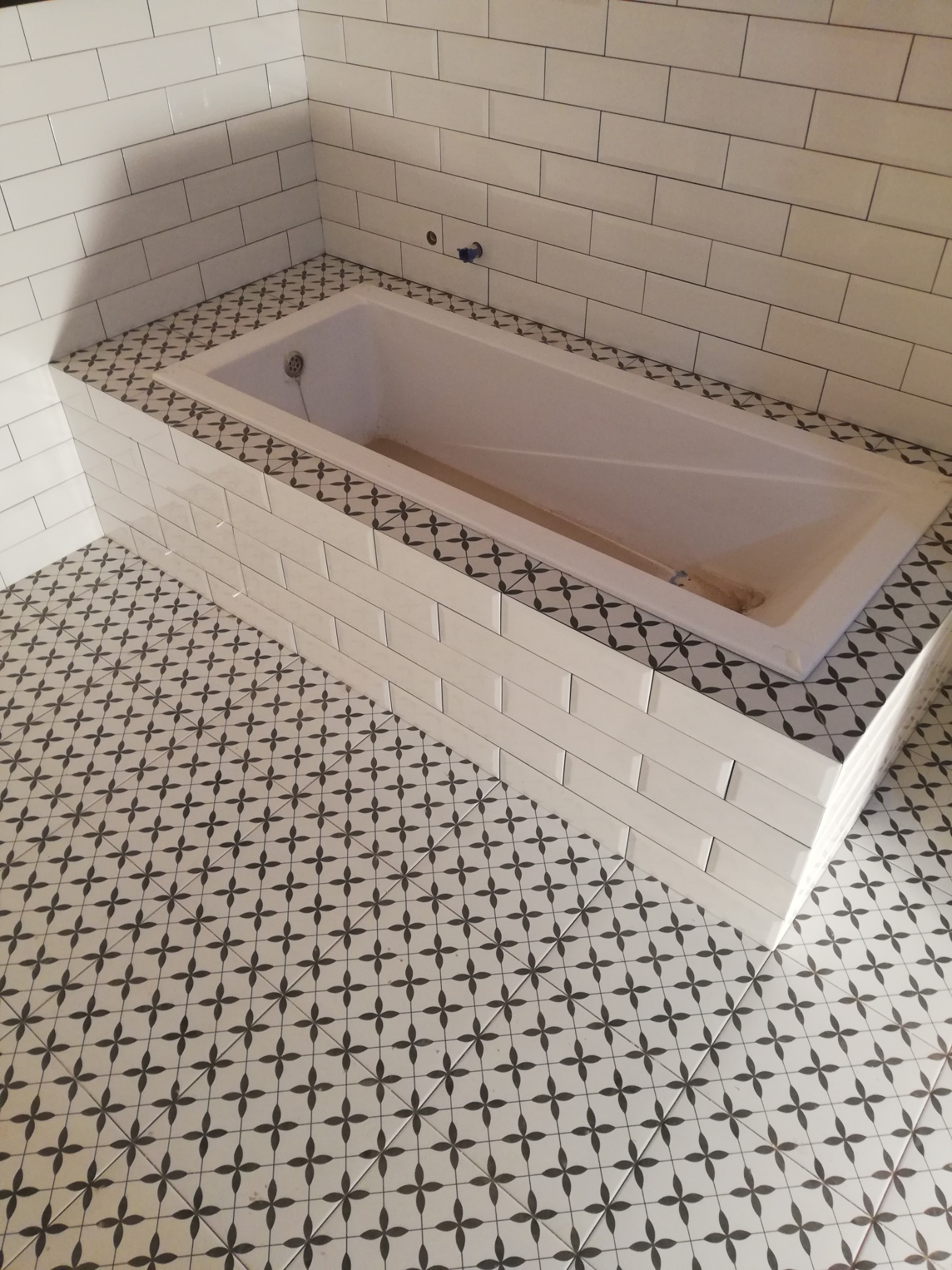 Biała wanna obudowana płytkami w łazience, z dekoracyjnymi płytkami podłogowymi w wzór czterolistnej koniczyny i białymi prostokątnymi płytkami na ścianach.