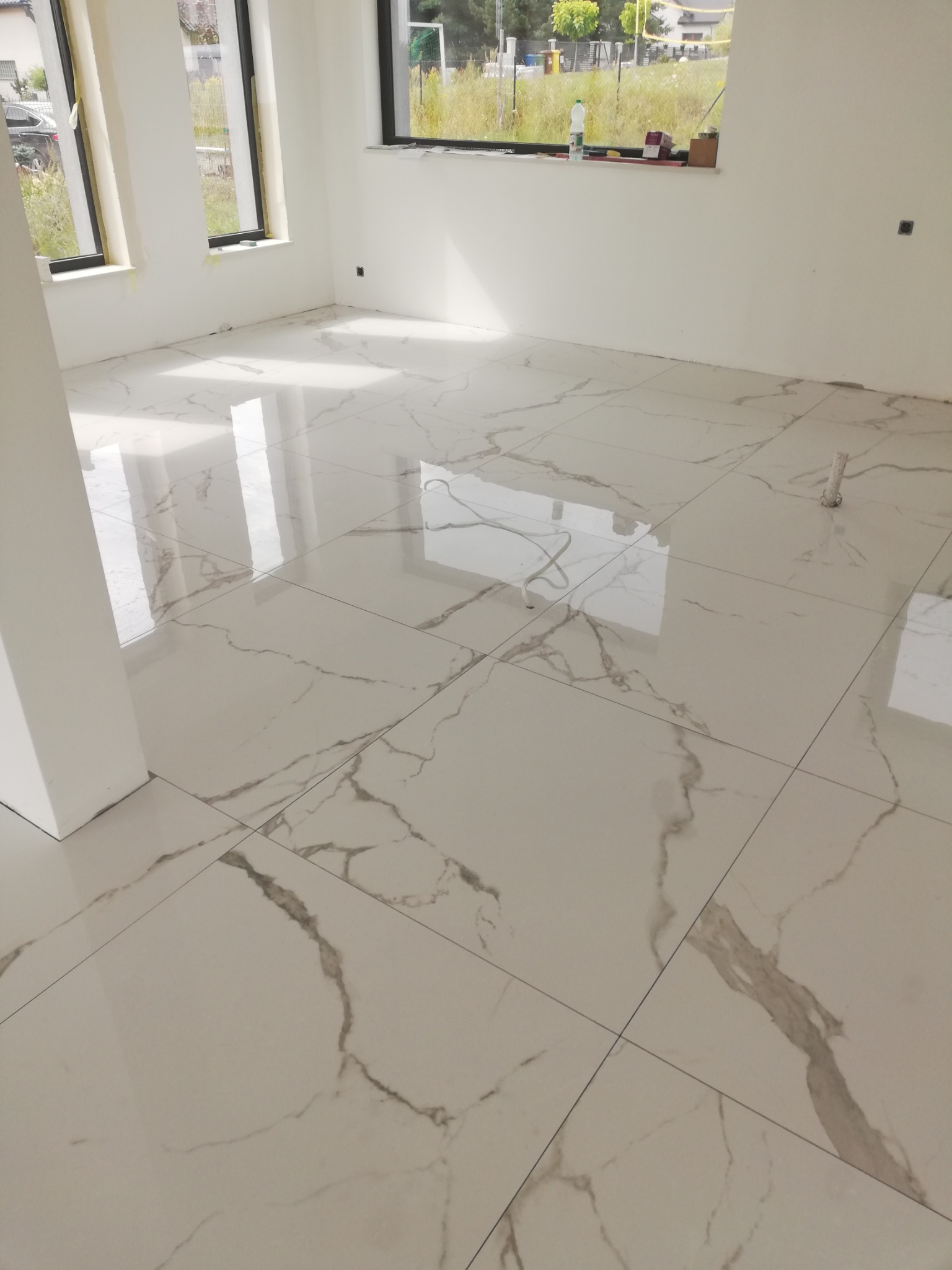 Wyłożona jasnymi, błyszczącymi płytkami podłoga w nowym, pustym pomieszczeniu z trzema oknami i białymi ścianami. Płytki imitują marmur z szarymi żyłami.