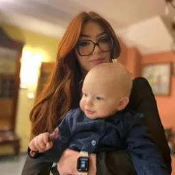 Młoda kobieta w okularach trzyma na rękach małe dziecko ubrane w granatową bluzkę. Kobieta ma długie, rude włosy i nosi smartwatch na nadgarstku.