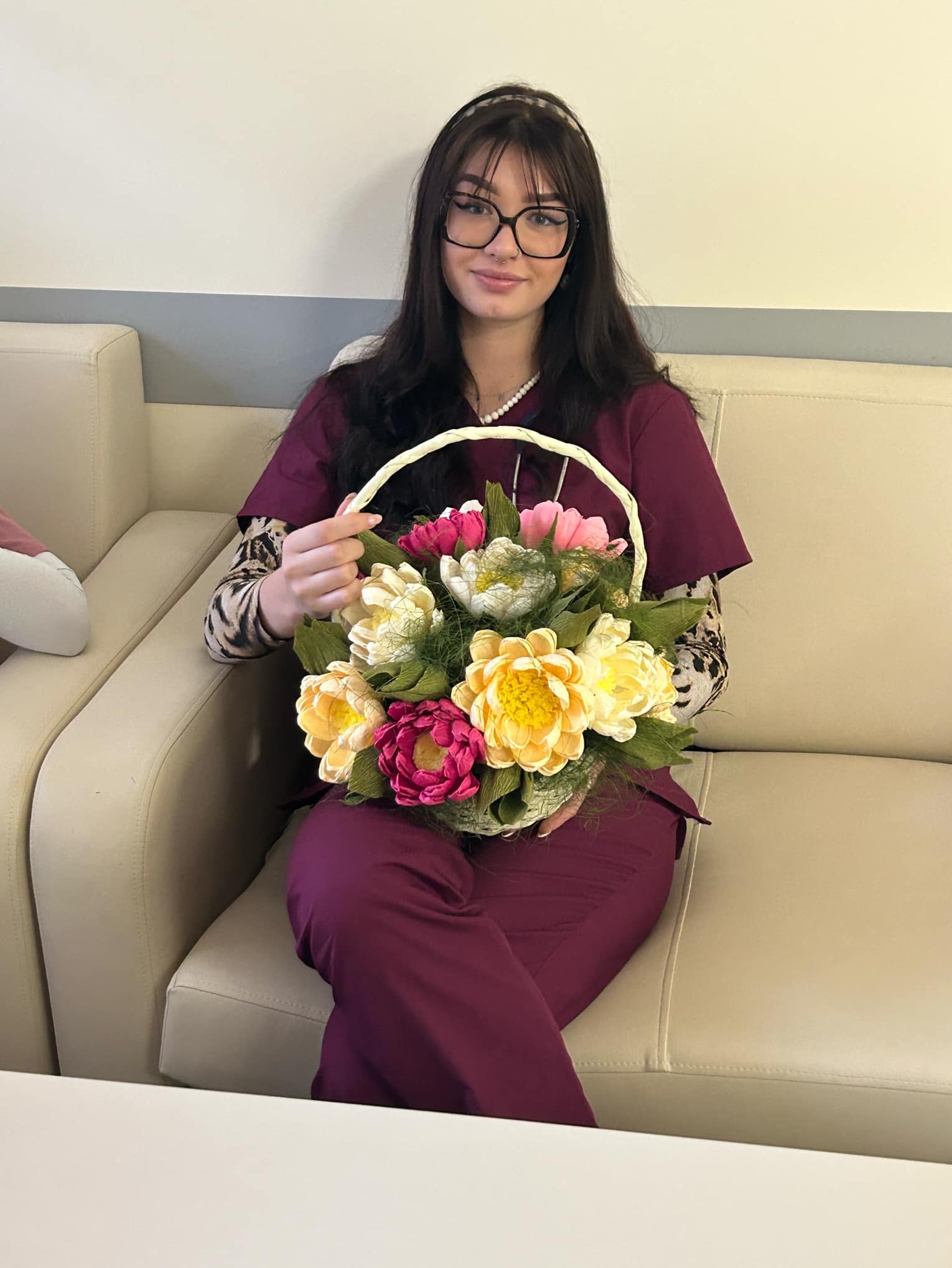 Uśmiechnięta kobieta w bordowym uniformie siedzi na beżowej sofie i trzyma kosz z kolorowymi, papierowymi kwiatami.