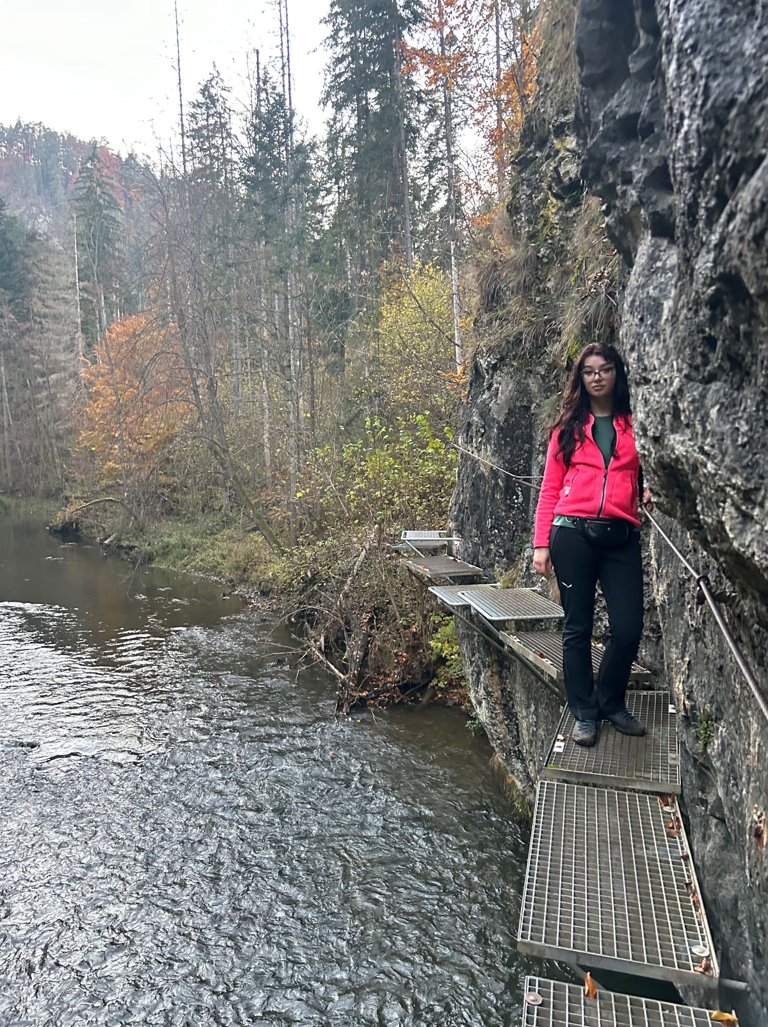 Kobieta w różowej kurtce i okularach idzie po metalowej kładce przymocowanej do skalnej ściany nad rzeką, w tle widoczny las.