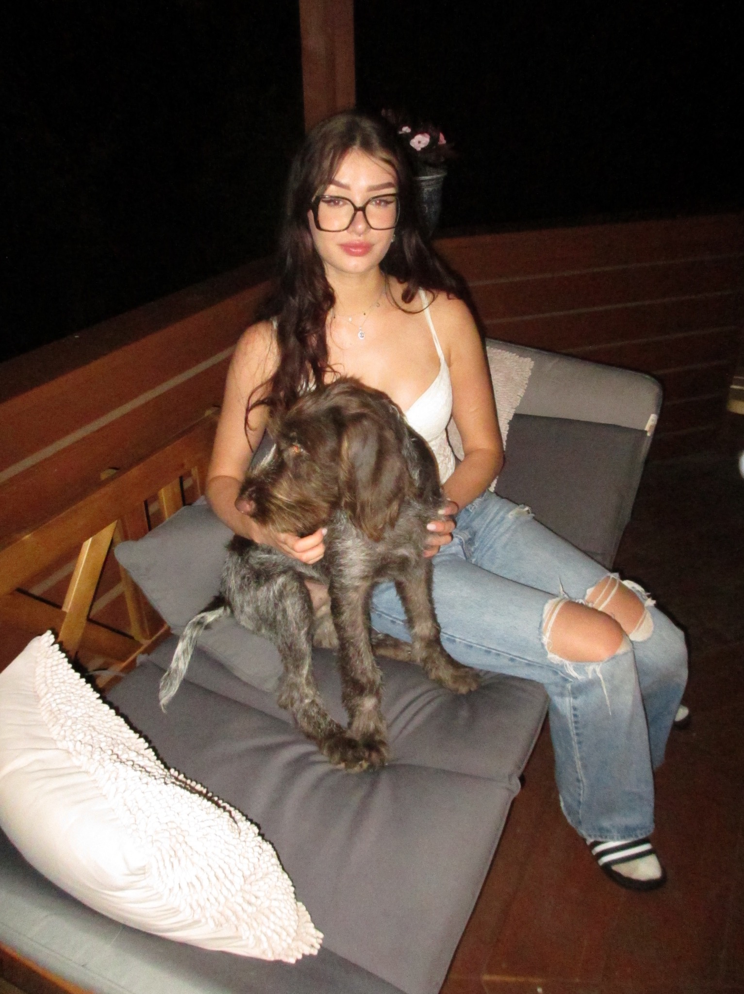 Młoda kobieta w okularach i podartych jeansach siedzi na leżaku na drewnianym tarasie, trzymając na kolanach szorstkowłosego psa w brązowo-szarym umaszczeniu, kwiaty w doniczce w tle.