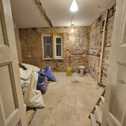 LIKIO - Wnętrze pokoju przed remontem: odsłonięta cegła na ścianach, dwa okna z różnymi ramami, worki z materiałami budowlanymi, stara toaleta i drabina.