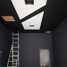 LIKIO - Czarno-biały sufit z geometrycznymi wzorami i otworami na oświetlenie, widoczna drabina i pomalowane na czarno ściany w pomieszczeniu w trakcie prac wykończeniowych.