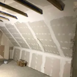 Poddasze w trakcie adaptacji: widoczne drewniane belki stropowe, ściany pokryte płytami gipsowo-kartonowymi z widocznymi łączeniami szpachlowymi, elementy instalacji elektrycznej oraz materiały...