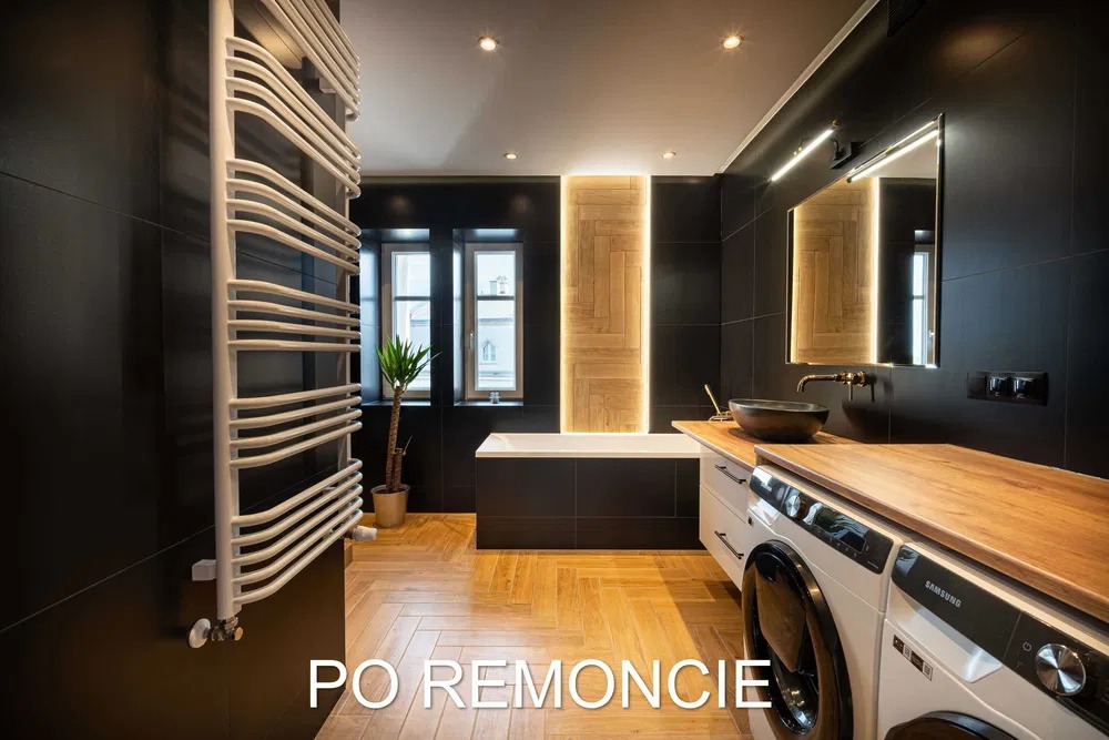 Nowoczesna łazienka z czarnymi płytkami, drewnianą podłogą ułożoną w jodełkę, grzejnikiem drabinkowym, umywalką na drewnianym blacie i pralką.