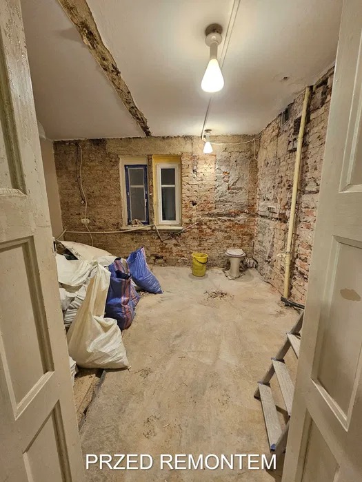 Wnętrze pokoju przed remontem: odsłonięta cegła na ścianach, dwa okna z różnymi ramami, worki z materiałami budowlanymi, stara toaleta i drabina.