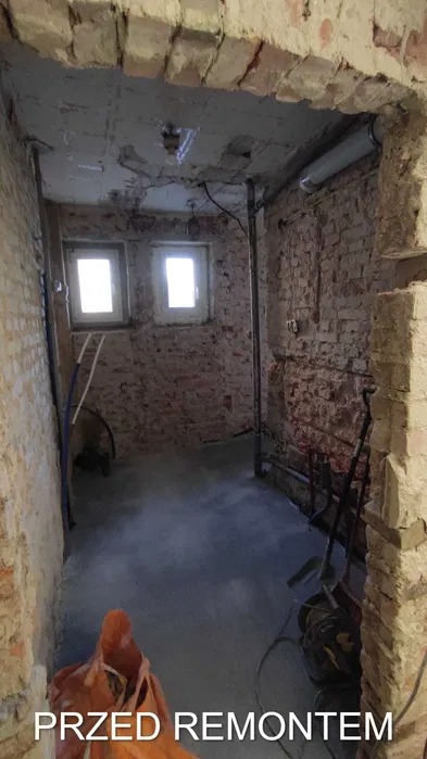 Wnętrze pomieszczenia z surowymi ścianami z cegły i betonową posadzką w trakcie prac przygotowawczych do remontu, widoczne okna i instalacje.
