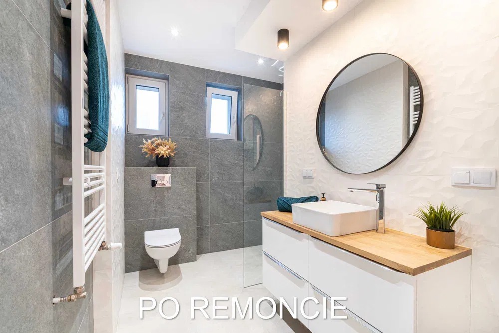 Nowoczesna łazienka po remoncie z szarymi płytkami, białymi meblami z drewnianym blatem i okrągłym lustrem.