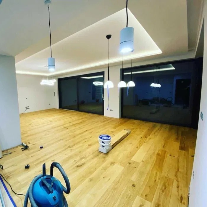Jasna, drewniana podłoga po ułożeniu, widoczny odkurzacz, puszka z farbą, narzędzia, duże okna przesuwne i nowoczesne oświetlenie sufitowe.
