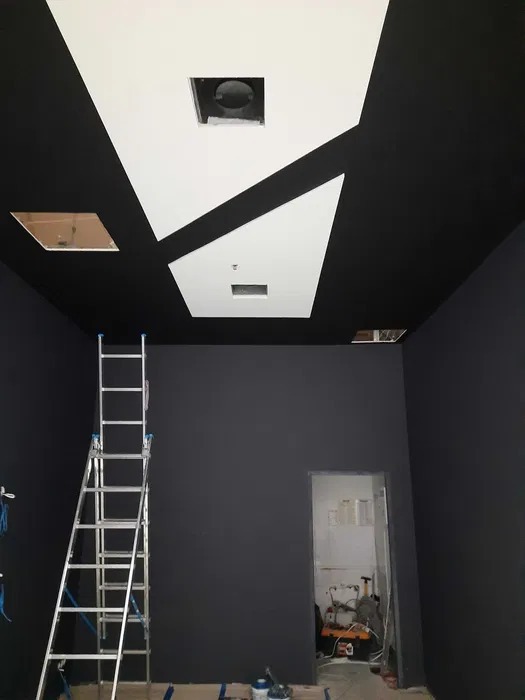 Czarno-biały sufit z geometrycznymi wzorami i otworami na oświetlenie, widoczna drabina i pomalowane na czarno ściany w pomieszczeniu w trakcie prac wykończeniowych.