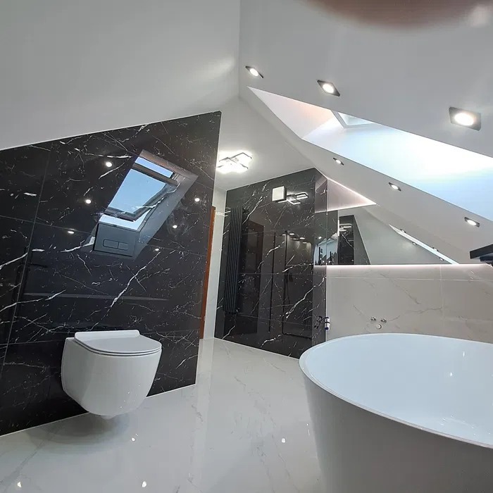 Nowoczesna łazienka na poddaszu z czarnymi płytkami imitującymi marmur na ścianach, białymi płytkami na podłodze, wiszącą toaletą i owalną wanną wolnostojącą, oświetlona punktowymi lampami...