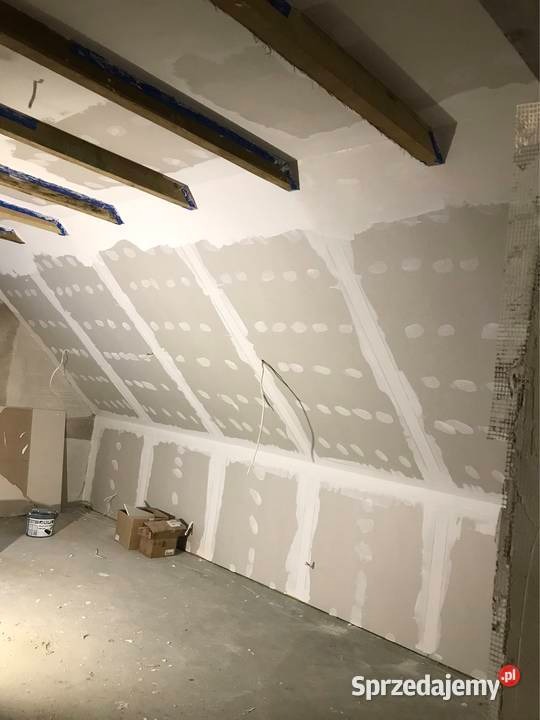 Poddasze w trakcie adaptacji: widoczne drewniane belki stropowe, ściany pokryte płytami gipsowo-kartonowymi z widocznymi łączeniami szpachlowymi, elementy instalacji elektrycznej oraz materiały...