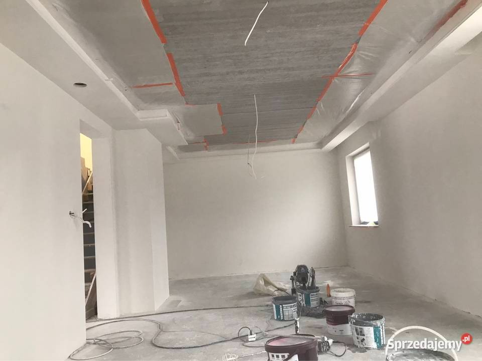 Surowe wnętrze w Krosno przed ostatecznym wykończeniem: świeżo pomalowane ściany na biało, widoczne puszki z farbą i narzędzia malarskie na podłodze, sufit częściowo zabezpieczony folią malarską.