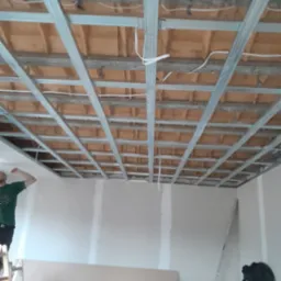 Montaż metalowego stelażu sufitu podwieszanego z widoczną izolacją wełną mineralną oraz częściowo wykończone ściany gipsowe, w tle pracownik na drabinie.