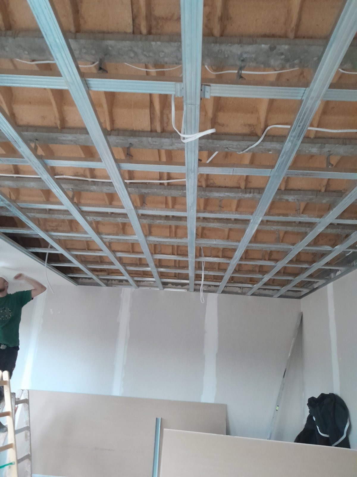 Montaż metalowego stelażu sufitu podwieszanego z widoczną izolacją wełną mineralną oraz częściowo wykończone ściany gipsowe, w tle pracownik na drabinie.