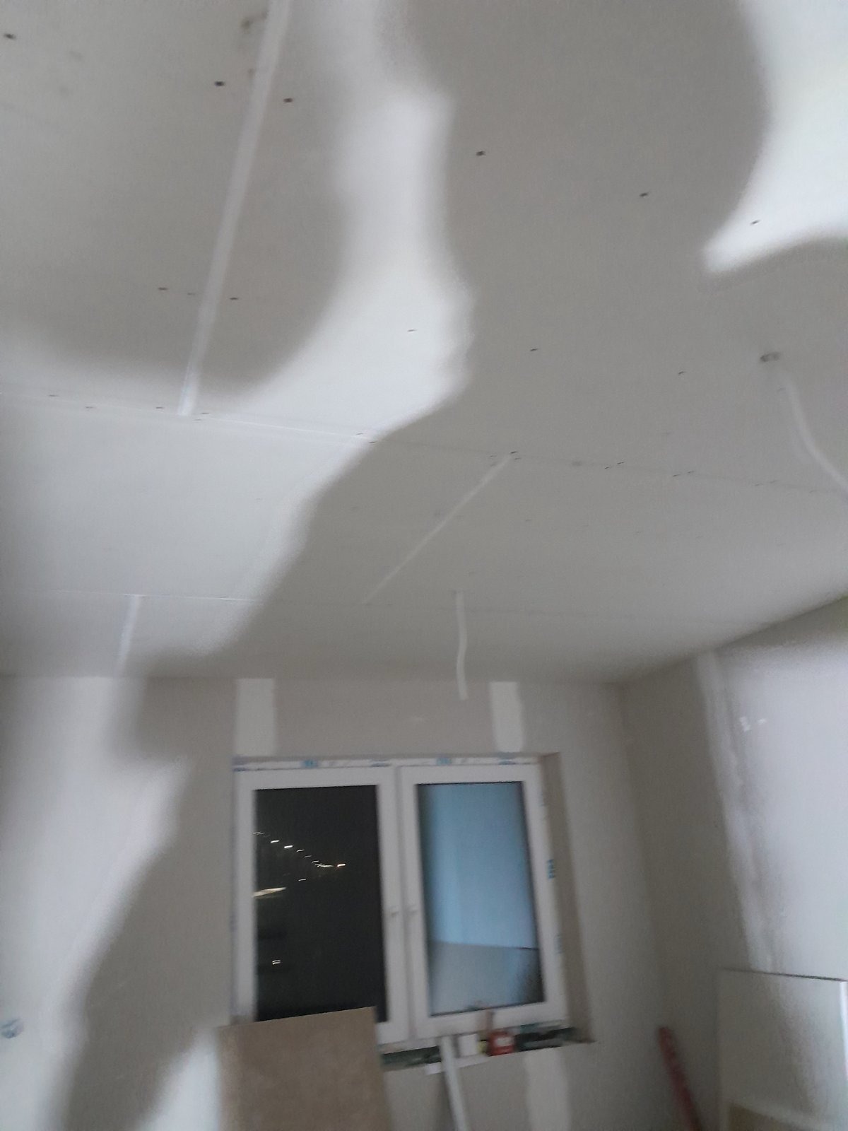 Częściowo wykończone wnętrze z zamontowanymi płytami kartonowo-gipsowymi na suficie, widoczne łączenia płyt i cień osoby robiącej zdjęcie.