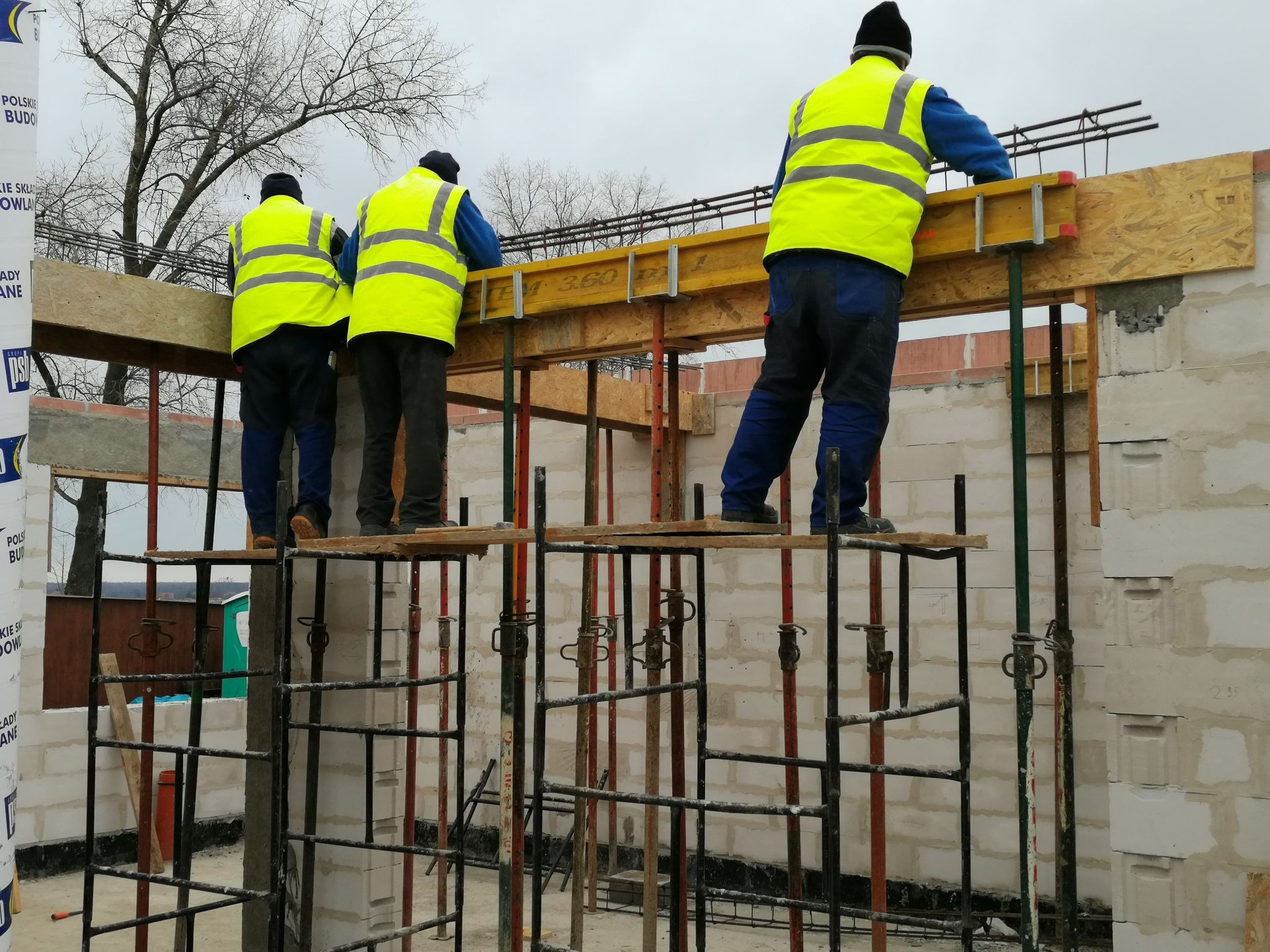 Trzech budowlańców w żółtych kamizelkach montuje szalunek i zbrojenie na budowie ściany z białych bloczków, widoczne podpory szalunkowe i fragment rusztowania.