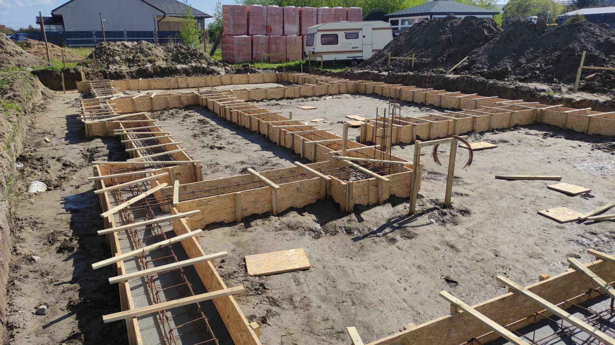 Przygotowany szalunek pod fundament domu jednorodzinnego z widocznym zbrojeniem, na drugim planie sterta cegieł i przyczepa kempingowa.
