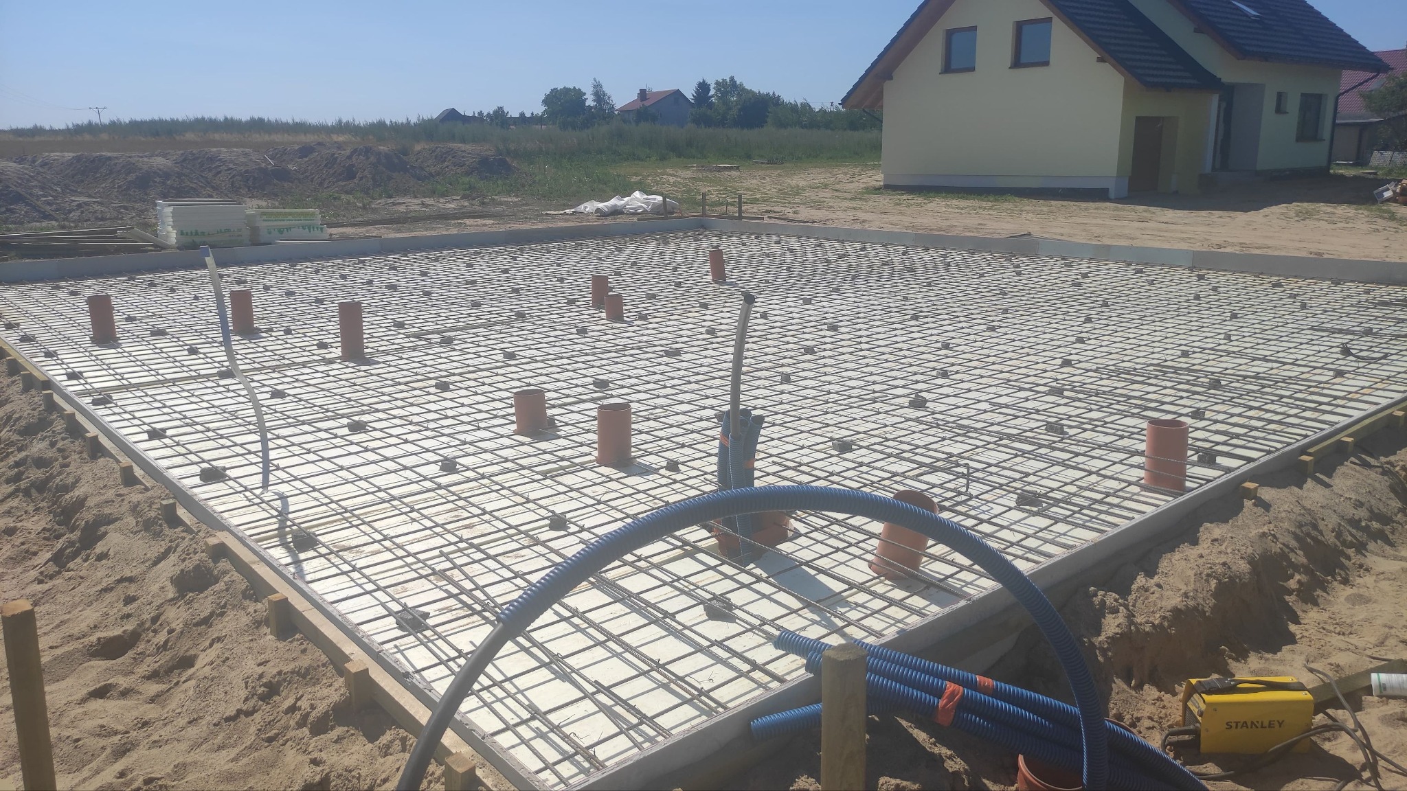 Zbrojenie płyty fundamentowej na białej izolacji, widoczne pionowe rury i niebieskie peszle, w tle żółty dom i wykop.