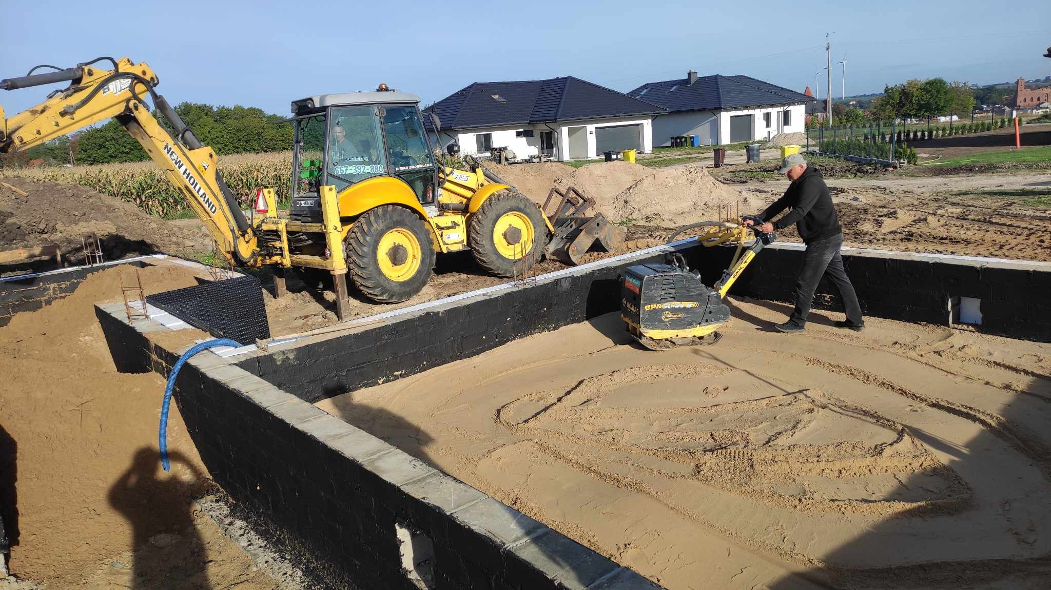 Wykop pod fundamenty domu jednorodzinnego z izolacją, ubijany piasek zagęszczarką wibracyjną, w tle żółta koparko-ładowarka New Holland i domy w budowie.