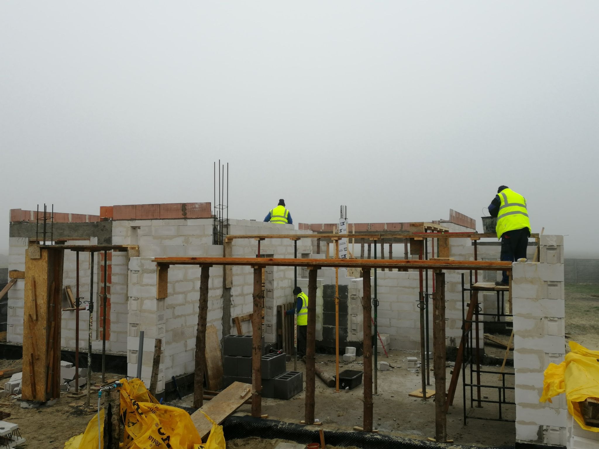 Wznoszenie ścian z bloczków betonu komórkowego na budowie domu jednorodzinnego, widoczni pracownicy w odblaskowych kamizelkach, drewniane stemple podtrzymujące konstrukcję, pochmurna pogoda.