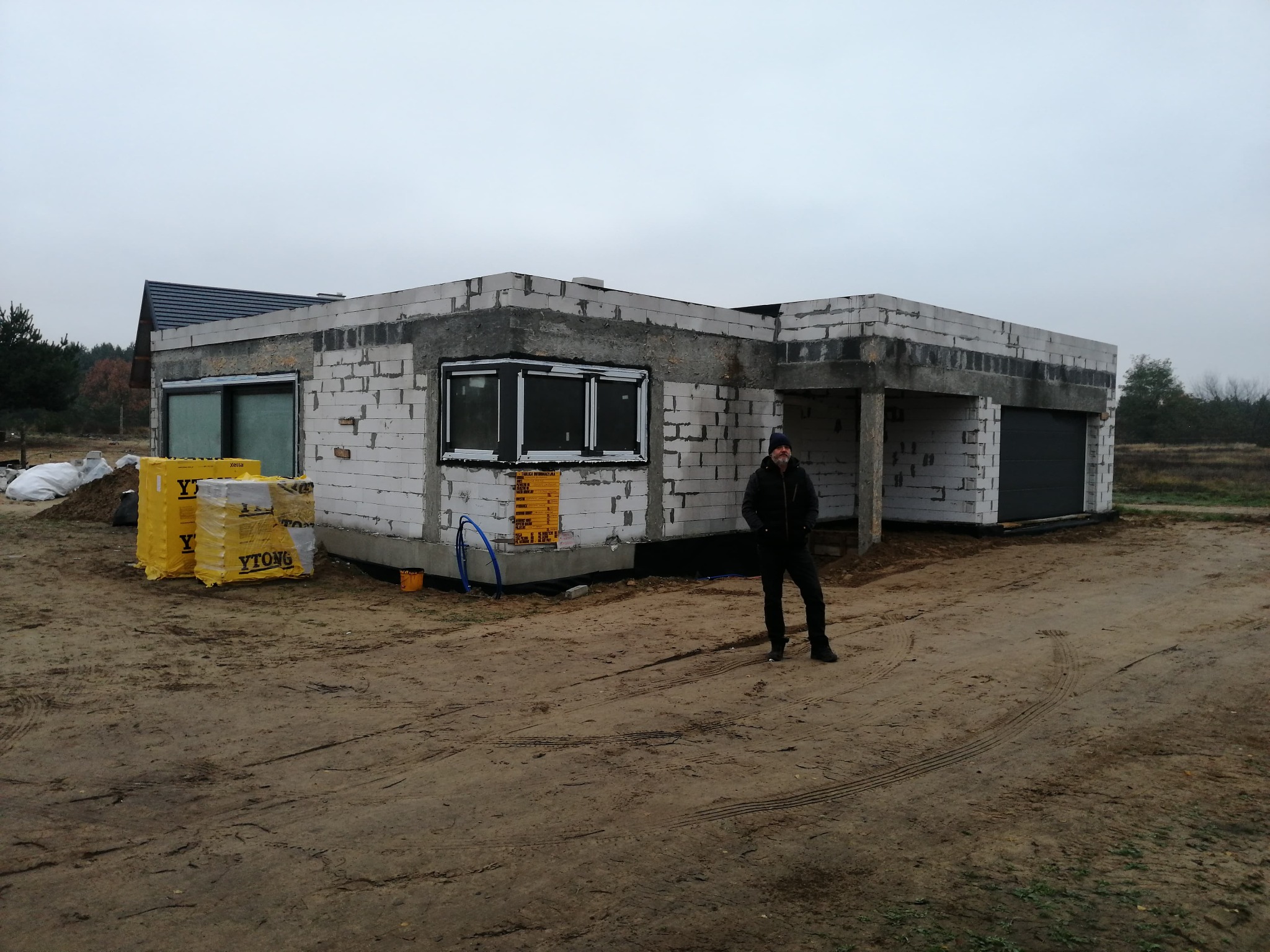 Niedokończony parterowy budynek z białych bloczków, z widocznymi oknami, otwartym garażem i stertą materiałów budowlanych Ytong na błotnistym terenie, z mężczyzną stojącym przed budynkiem.