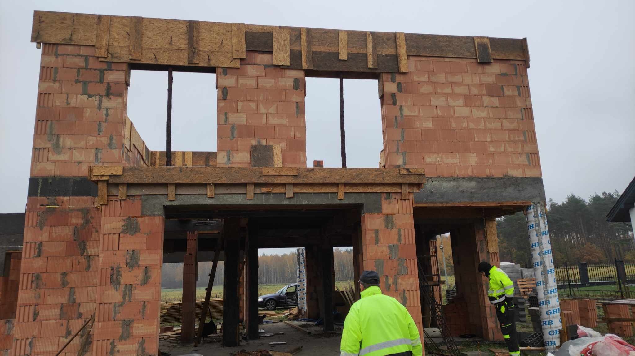 Wznoszenie ścian dwukondygnacyjnego budynku z czerwonej cegły dziurawki; widoczne otwory okienne i drzwiowe, drewniane szalunki nadproży, pracownik w odblaskowej kurtce, samochód w tle.