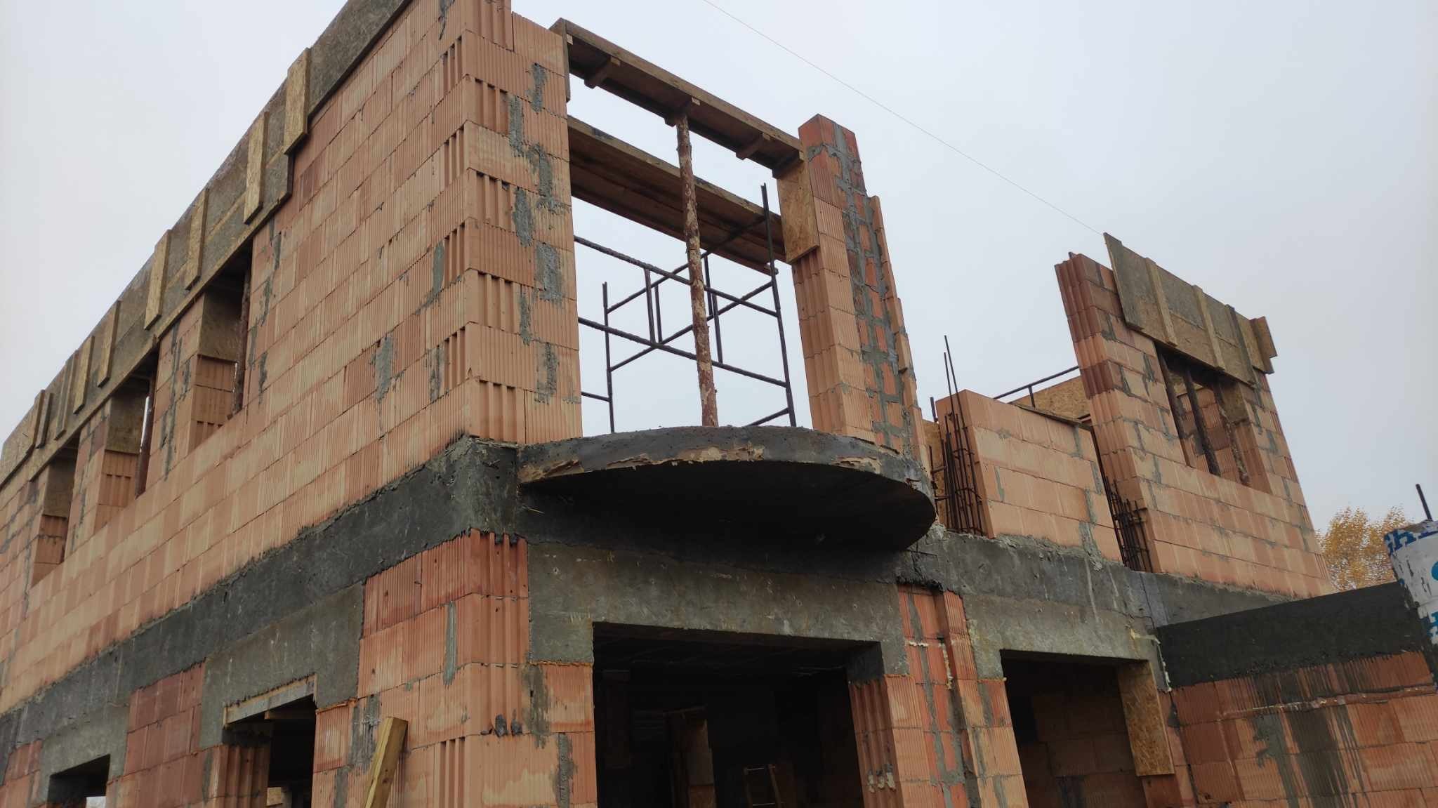 Wznoszenie dwukondygnacyjnego budynku z czerwonej ceramiki, widoczne elementy szalunków i rusztowania, zarys balkonu na pierwszym piętrze.