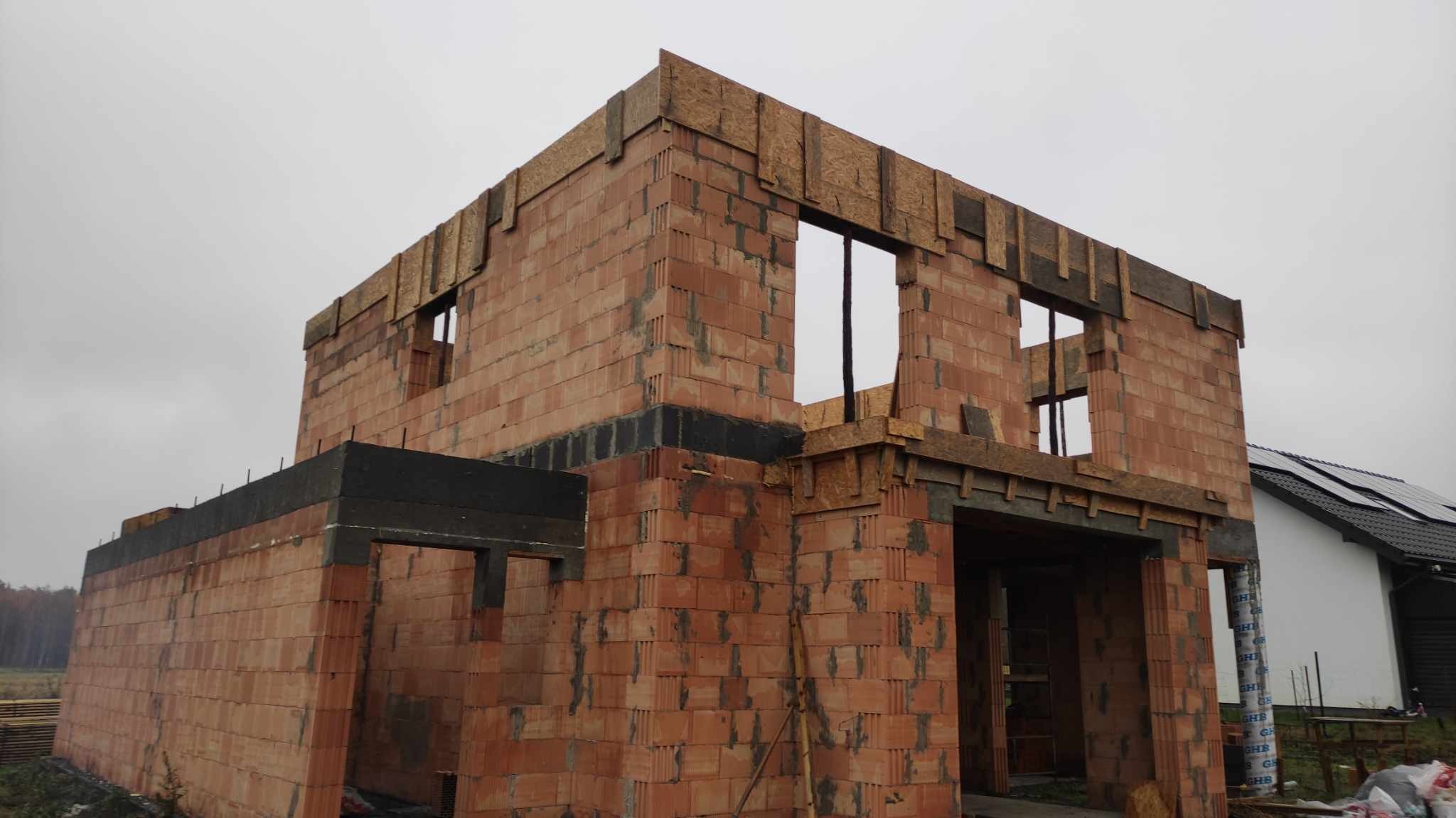 Dwukondygnacyjny dom w stanie surowym otwartym, ściany z czerwonej cegły ceramicznej, widoczne nadproża okienne i drzwiowe z drewnianych desek, ocieplone fundamenty, szary, pochmurny dzień.
