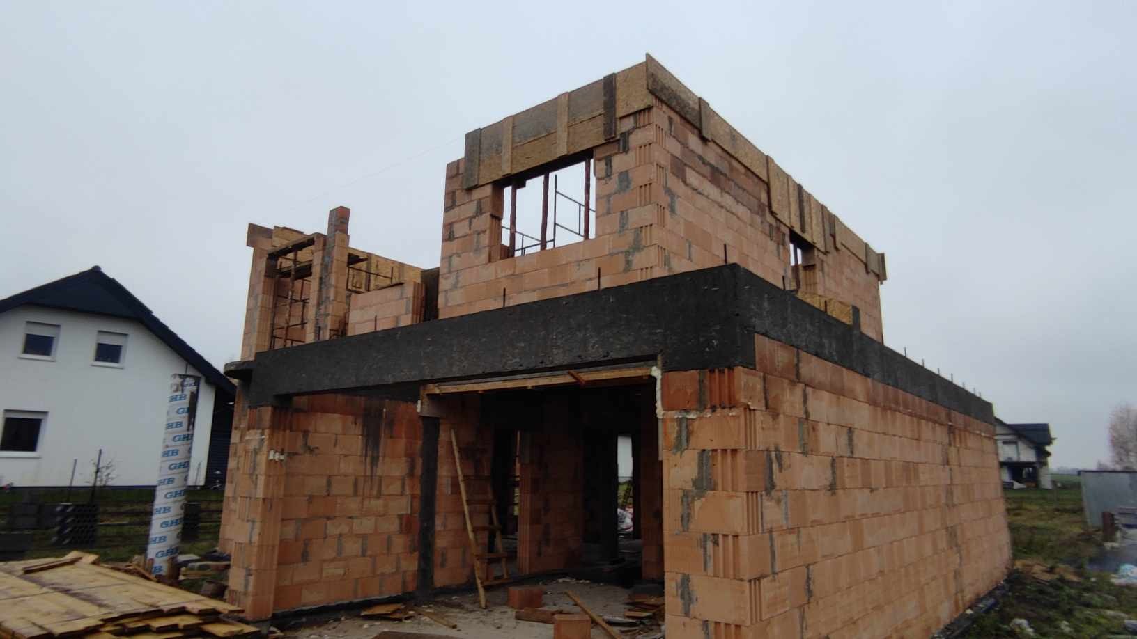 Surowy stan dwukondygnacyjnego domu w trakcie budowy z czerwonej cegły, widoczna czarna izolacja, drewniane elementy szalunkowe i drabina oparta o ścianę, w tle zabudowa jednorodzinna i pochmurne...
