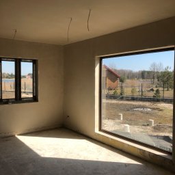 Damian Kaminski - Surowe wnętrze domu w trakcie wykańczania, ściany po wstępnym wyrównaniu, widoczne wystające przewody elektryczne z sufitu, duże okno z widokiem na jezioro i las.
