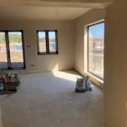 Wnętrze domu w stanie deweloperskim po tynkowaniu maszynowym, widoczne worki z tynkiem gipsowym Alfa Leikki Plus, okna z widokiem na jezioro, podłoga przygotowana do dalszych prac.