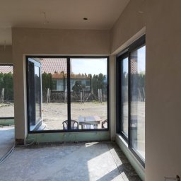 Damian Kaminski - Surowe ściany w nowym domu z dużymi oknami, widoczne przewody elektryczne wystające ze ścian, podłoga w stanie surowym, widok na ogród przez okna.