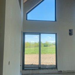 Damian Kaminski - Wnętrze w stanie deweloperskim z oknami na zewnątrz, ściany po nałożeniu gładzi, widoczne niedoskonałości podłoża i brak wykończenia podłogi.