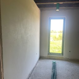 Damian Kaminski - Świeżo otynkowana ściana w surowym wnętrzu z widokiem na zewnątrz przez okno; na podłodze leżą zielone rury wentylacyjne.