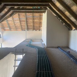 Damian Kaminski - Poddasze w trakcie prac wykończeniowych, widoczne surowe drewniane krokwie, ściany pokryte szarą warstwą, prawdopodobnie tynkiem, oraz rury instalacyjne ułożone na podłodze.